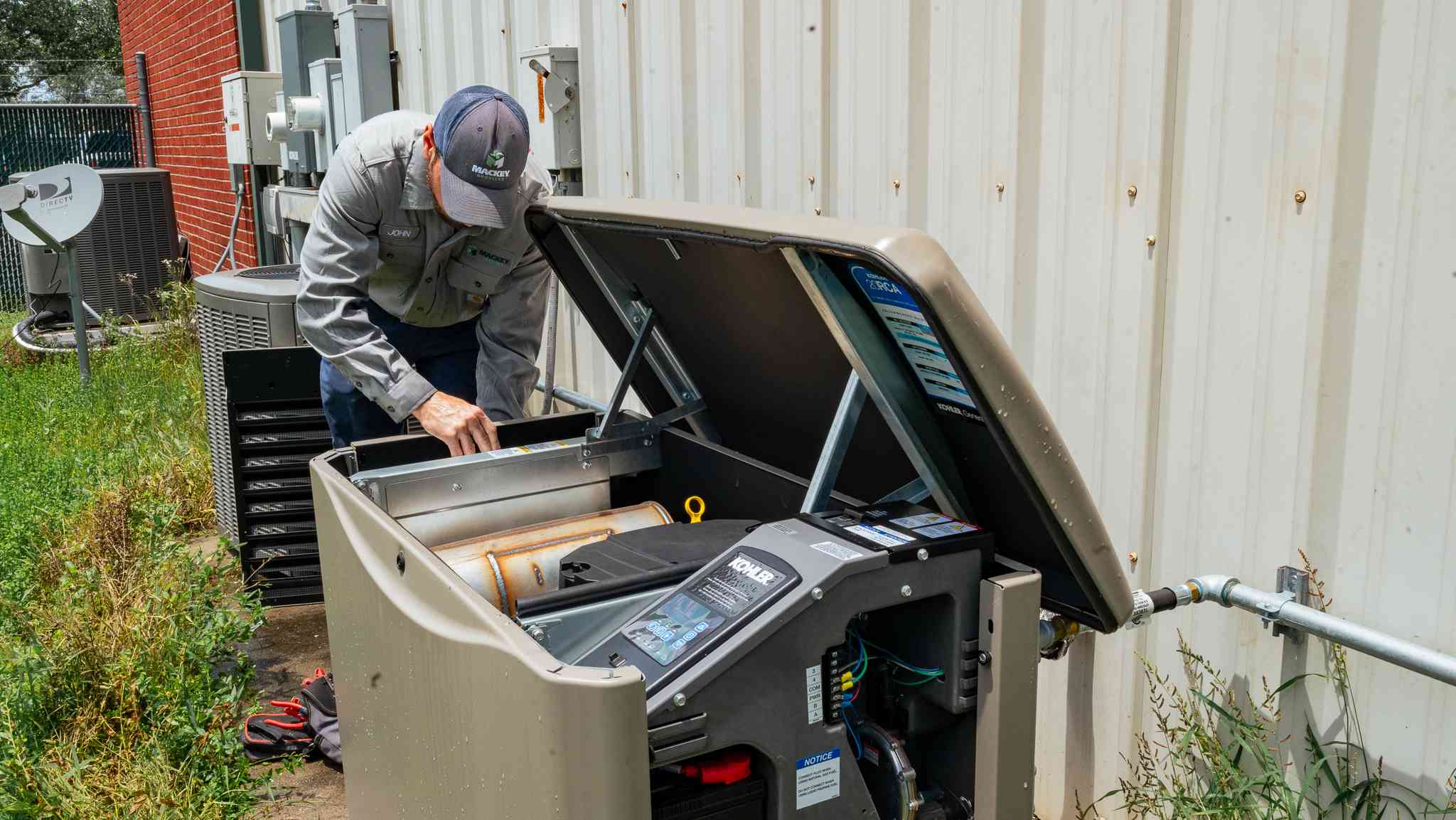 Technician in work uniform inspecting an open outdoor generator next to a building wall with grass and utility equipment nearby.