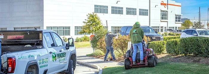 Two workers maintaining commercial landscaping near a white building; one is riding a lawn mower on grass while the other uses a tool near shrubs.