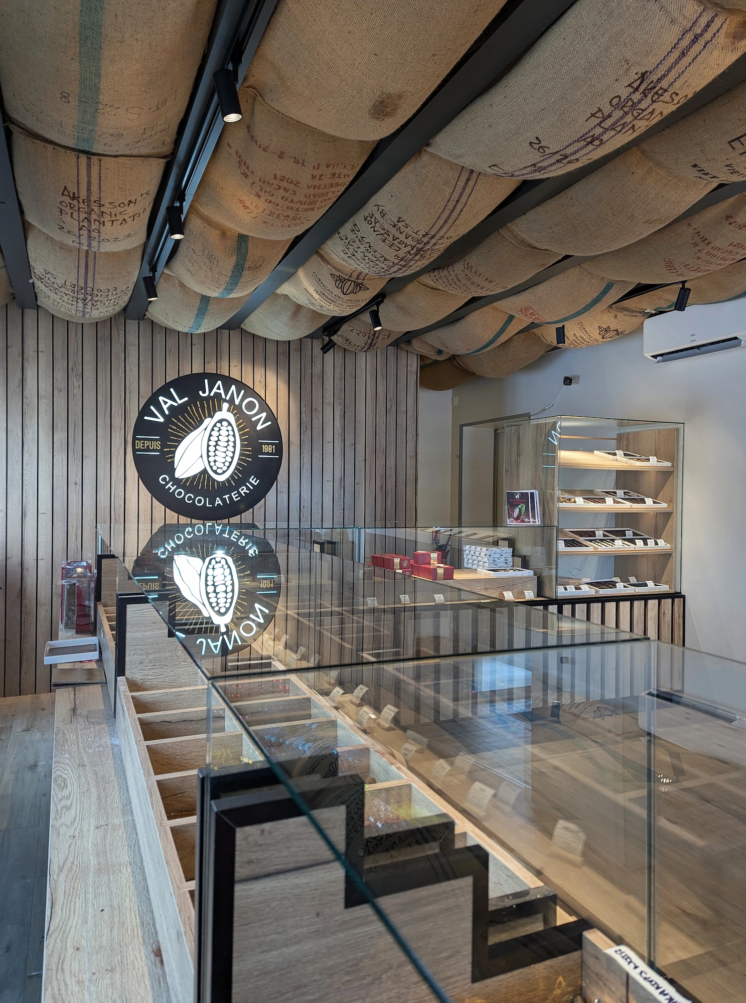 Comptoir en bois clair et présentation des produits dans la chocolaterie Val Janon, avec le logo mural en arrière-plan.
