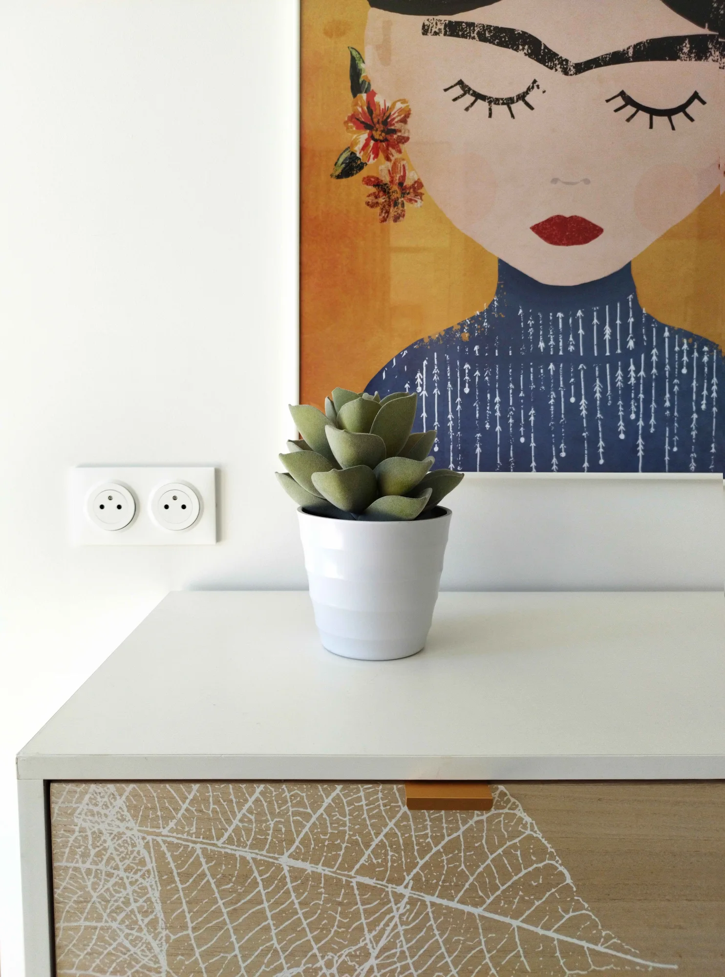 Détail décoratif : plante posée sur un meuble texturé sous un tableau illustré.