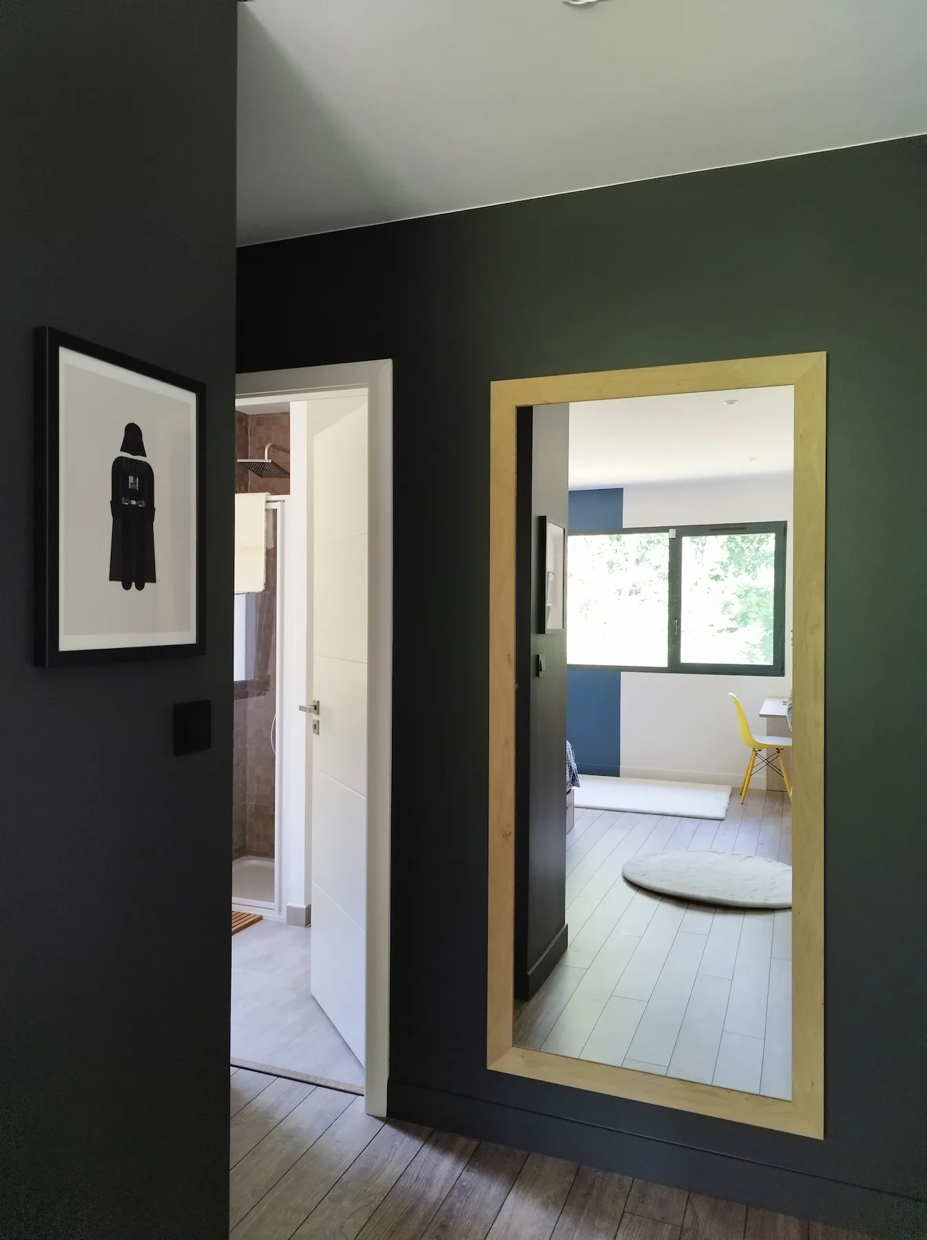 Vue d’un couloir sombre révélant un grand miroir à cadre bois clair, reflétant la chambre lumineuse attenante.