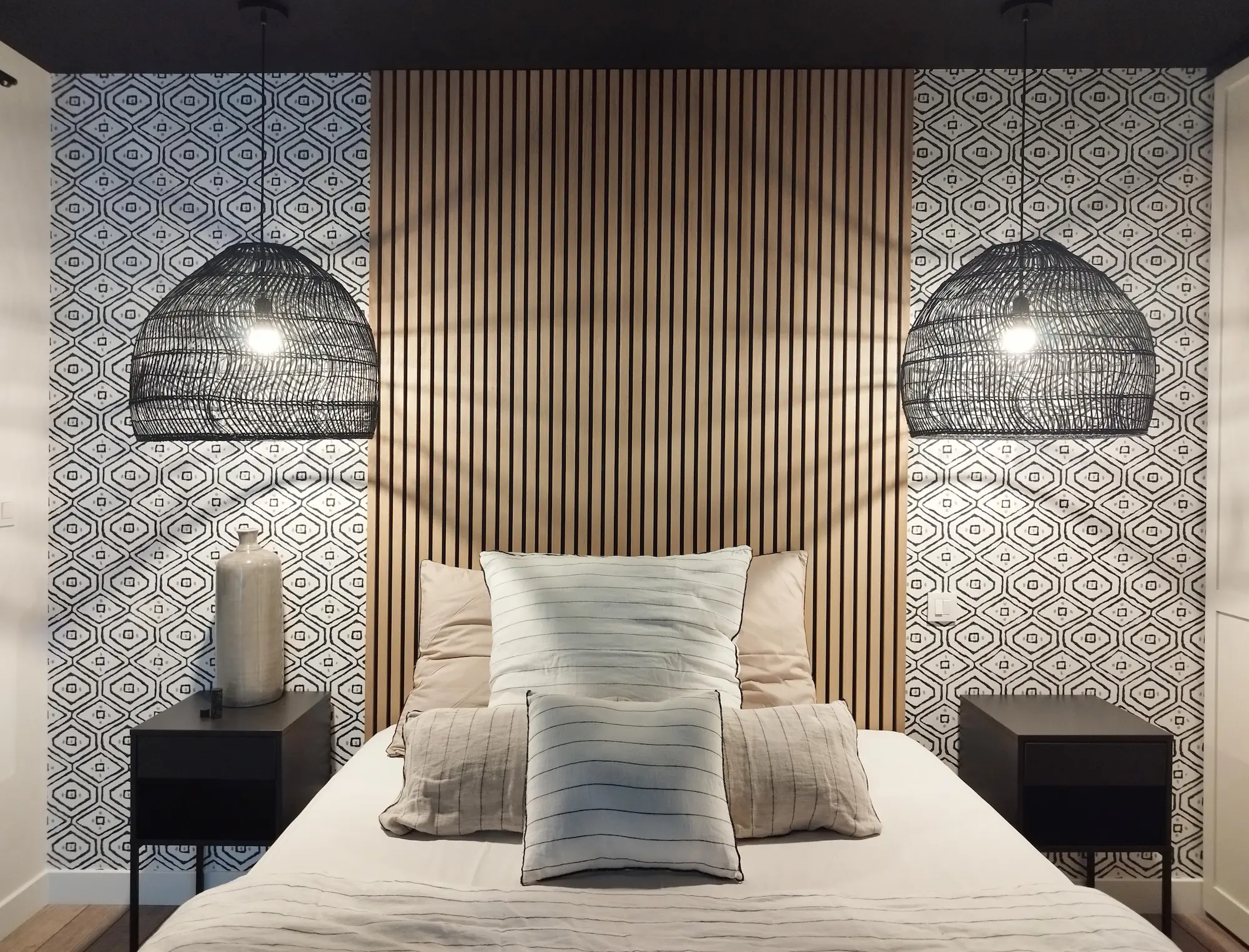 Tête de lit symétrique en tasseaux bois encadrée de papier peint graphique et suspension en rotin noir.