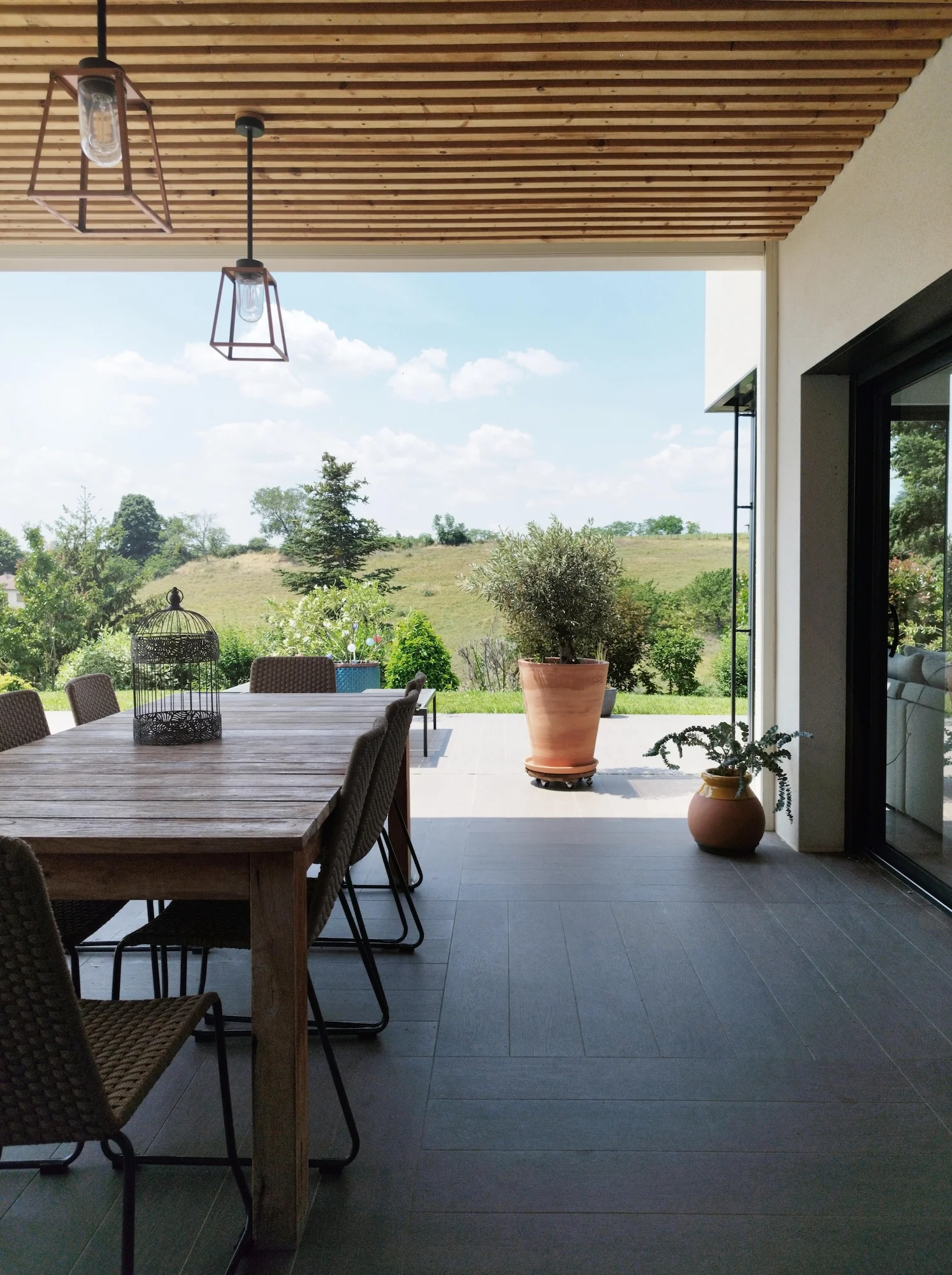 Terrasse couverte avec plafond bois et grande table extérieure, offrant une vue dégagée sur la campagne.