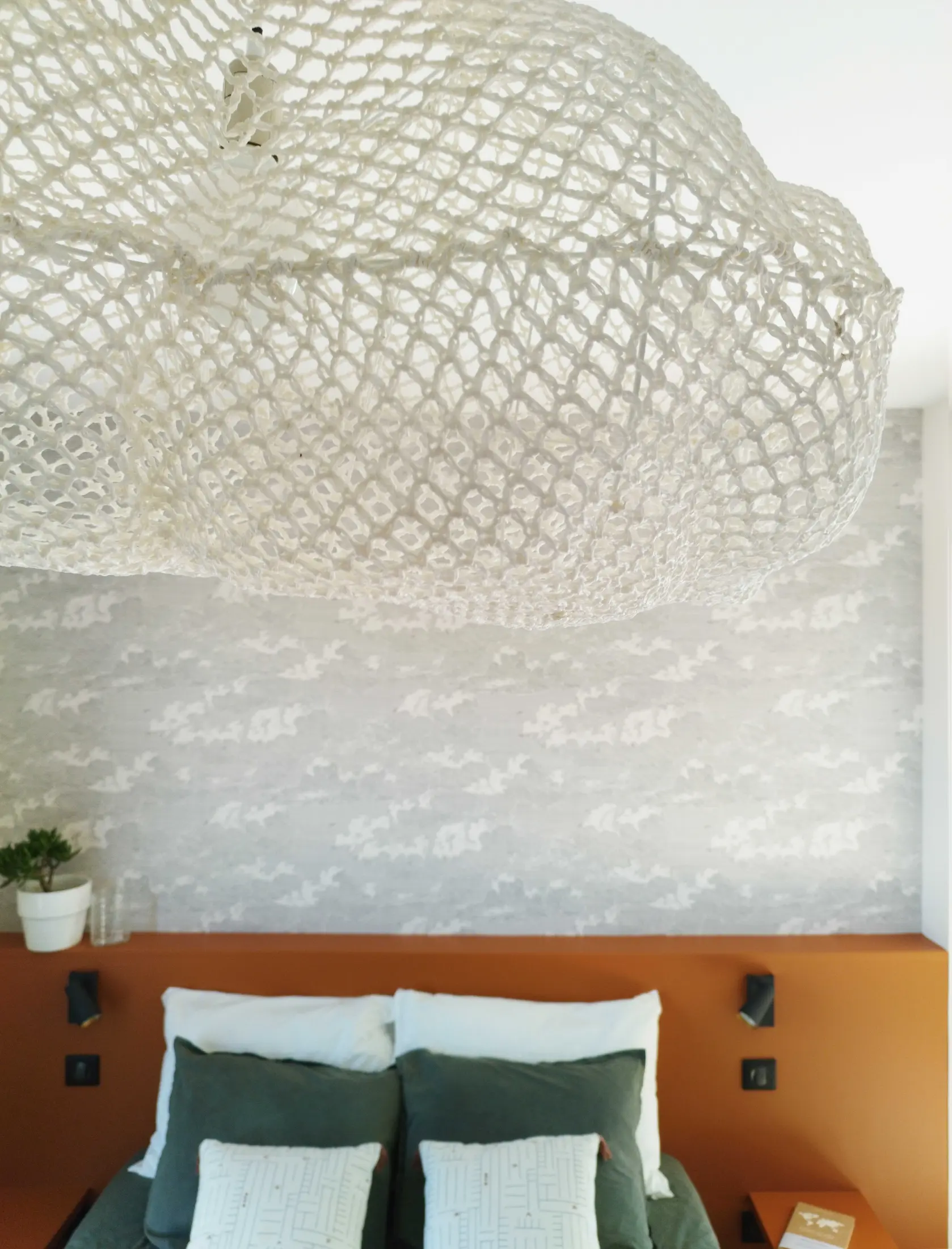 Vue en contre-plongée sur une suspension ajourée au-dessus d’une chambre contemporaine mêlant terracotta et papier peint gris.