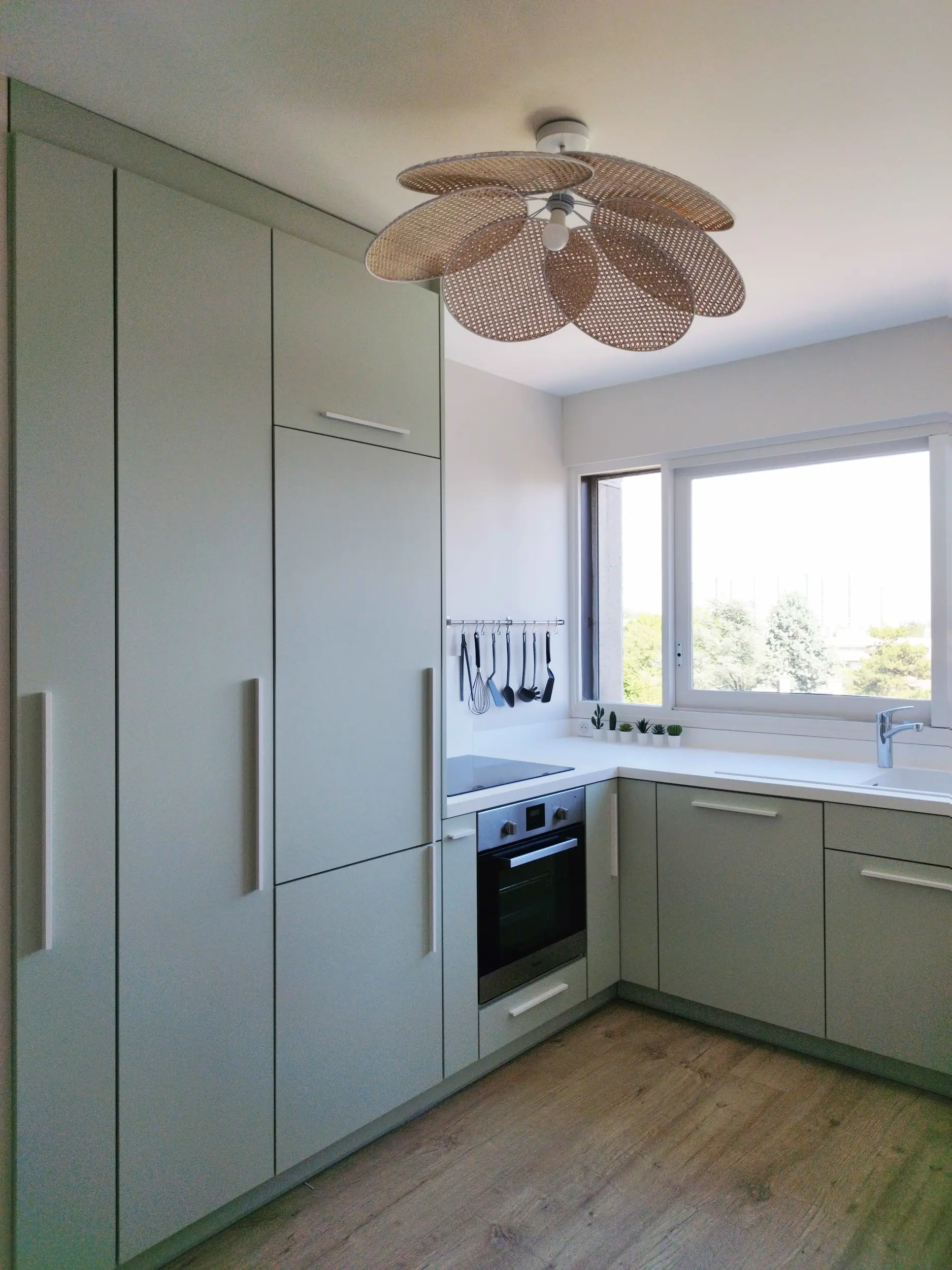 Vue d’ensemble de la cuisine sur-mesure, rénovée avec des façades verte et un luminaire en cannage.