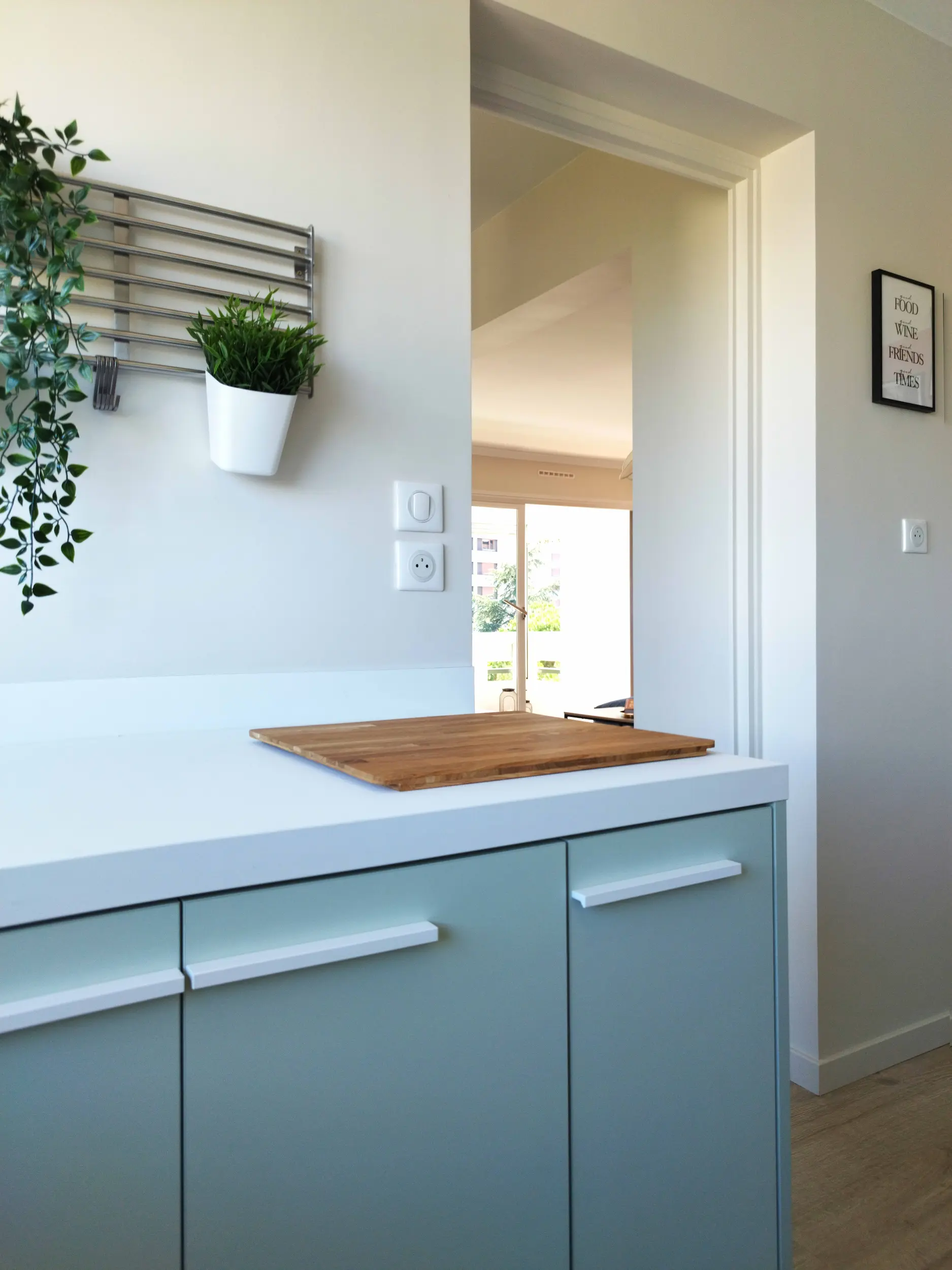 Plan de travail blanc et façades pastel dans la cuisine réaménagée, avec jardinière murale et vue traversante sur le séjour.