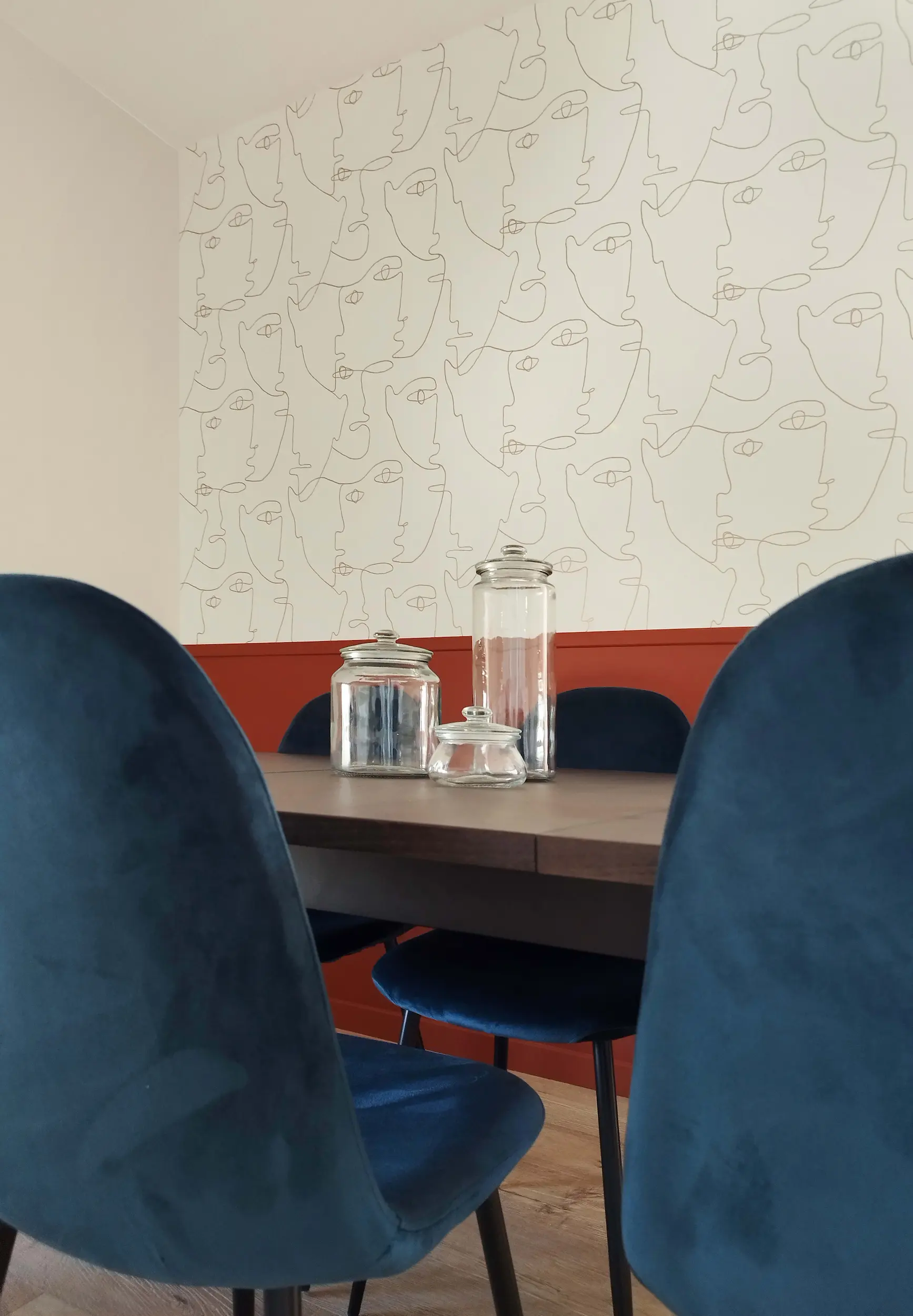 Salle à manger modernisée avec papier peint graphique façon visages, soubassement terracotta et chaises bleu nuit.