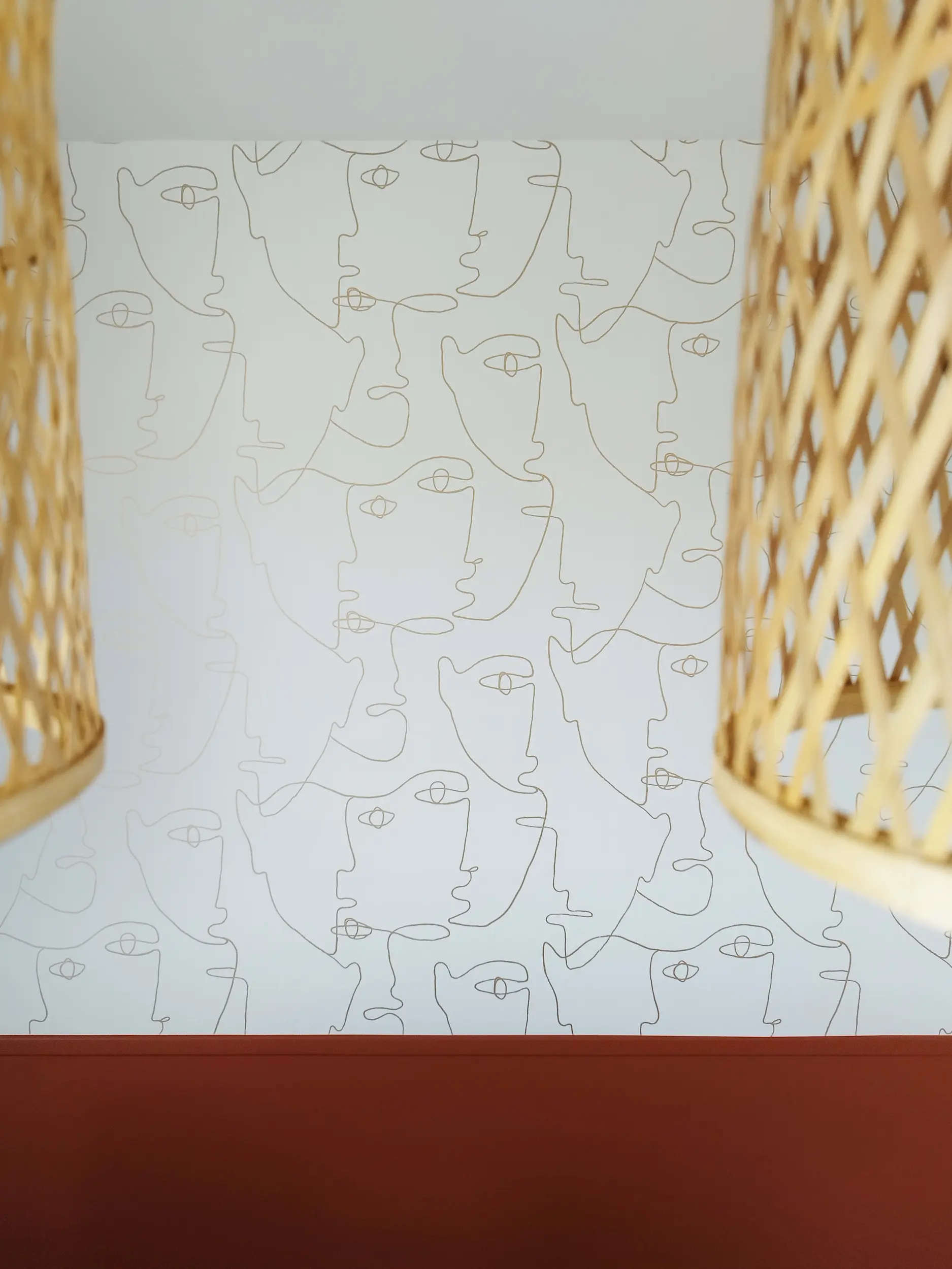 Papier peint graphique aux visages linéaires, encadré par deux suspensions en rotin dans la salle à manger.