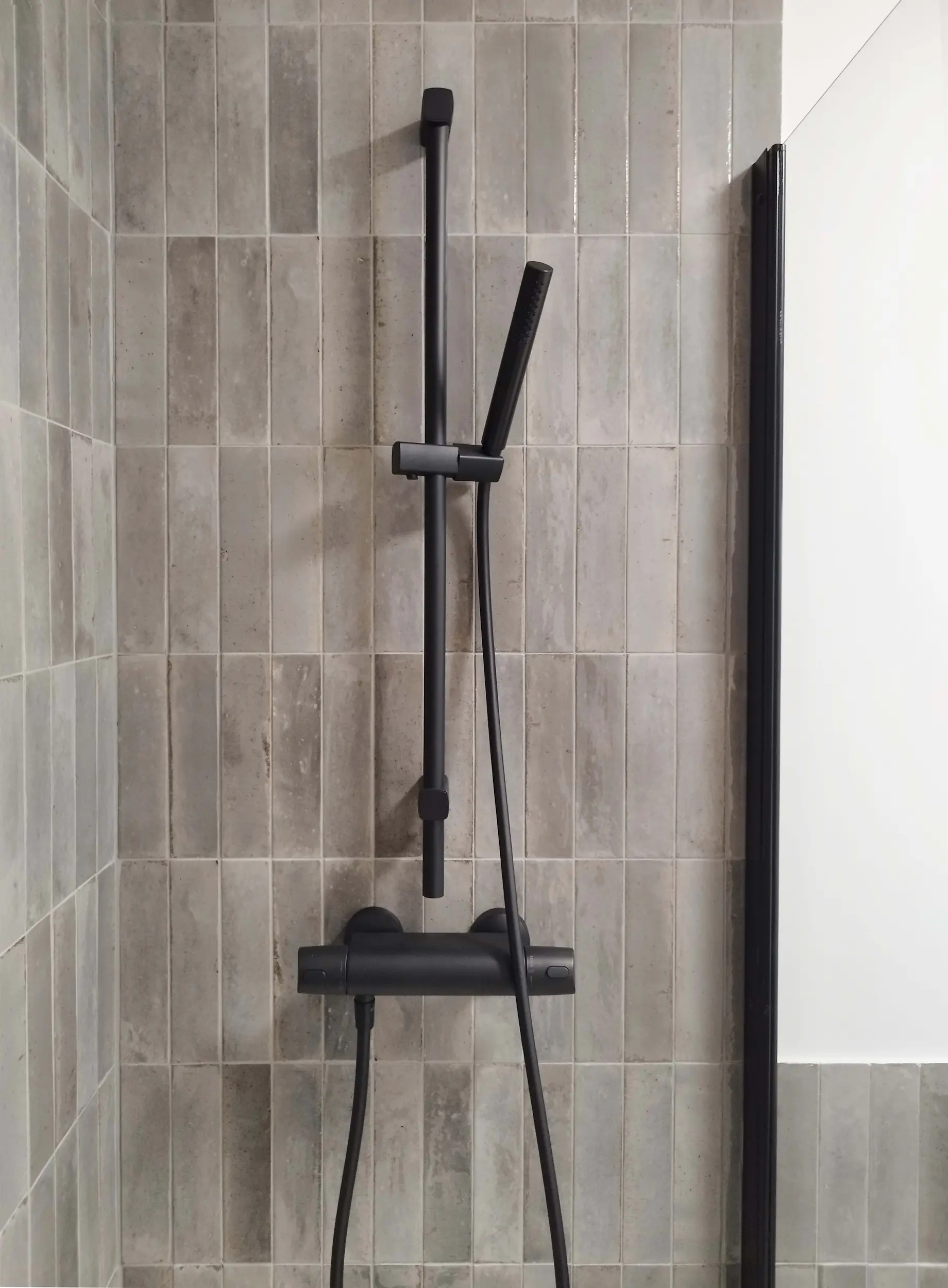 Barre de douche noire mate sur faïence verticale grise, dans la salle d’eau contemporaine.