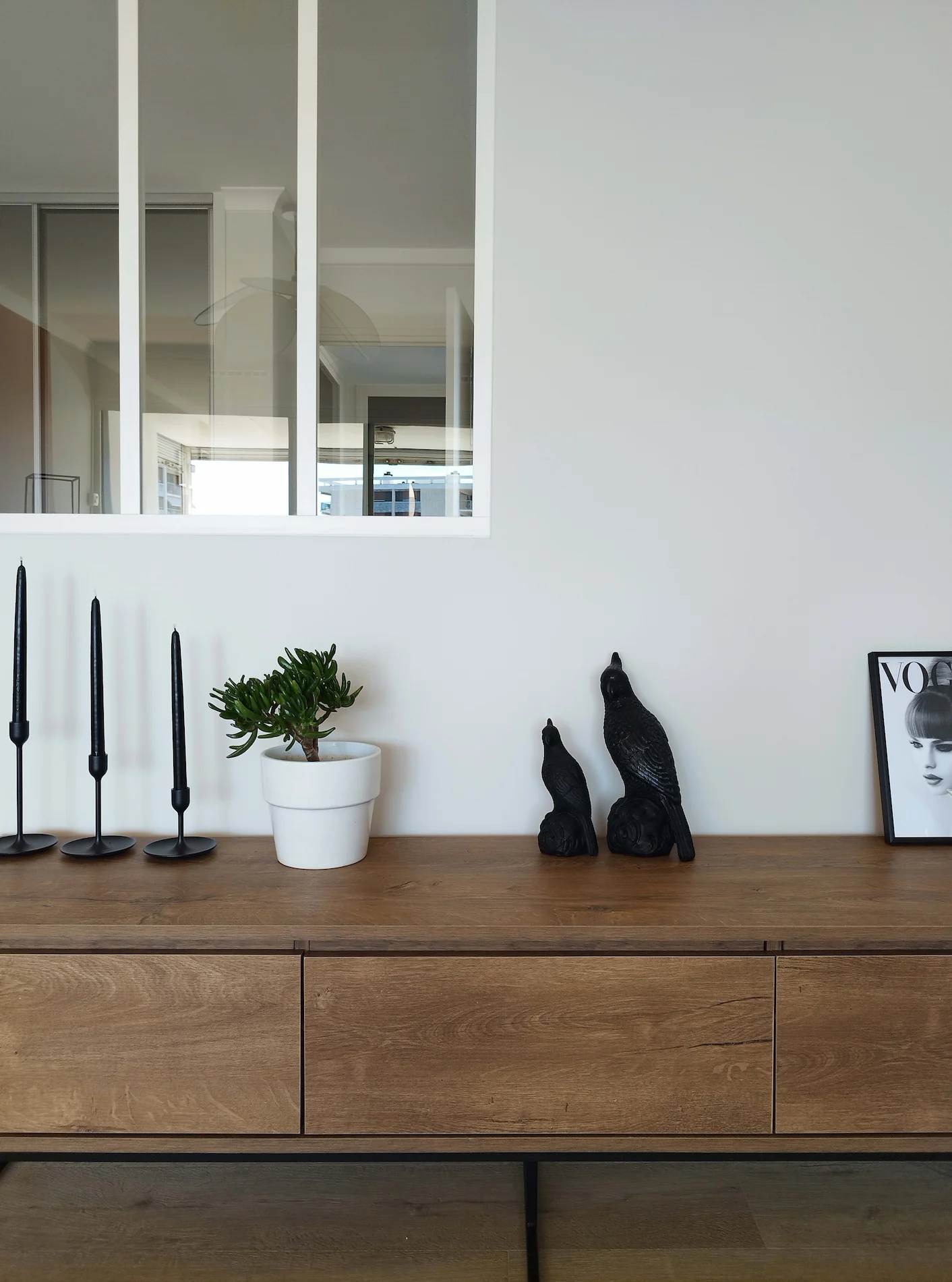 Console en bois avec objets décoratifs noirs et plante verte sous une verrière blanche, dans le séjour rénové de l’appartement.