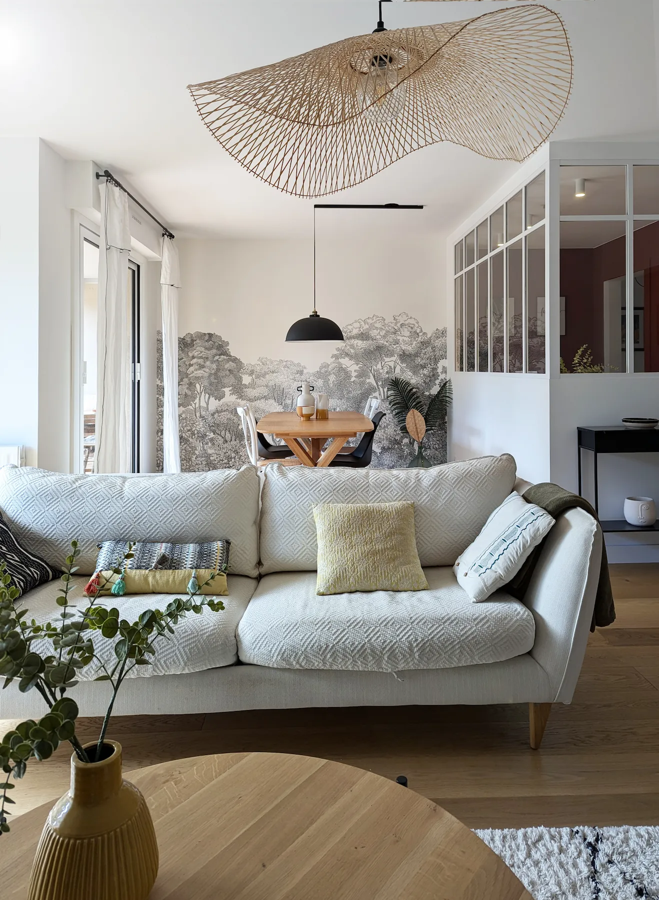 Salon d’un appartement rénové des années 70, avec canapé clair, papier peint panoramique végétal et ouverture sur la salle à manger.
