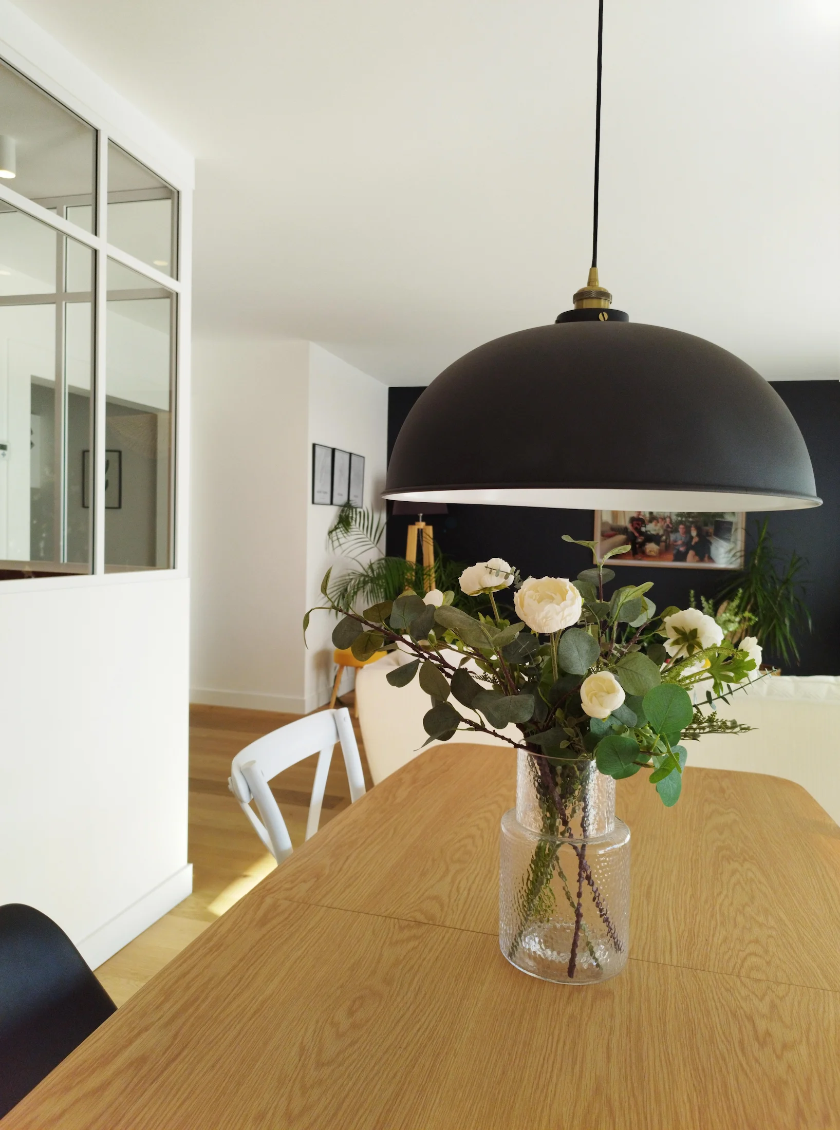 Suspension noire au-dessus d’une table en bois dans la salle à manger, avec verrière intérieure et décoration végétale.