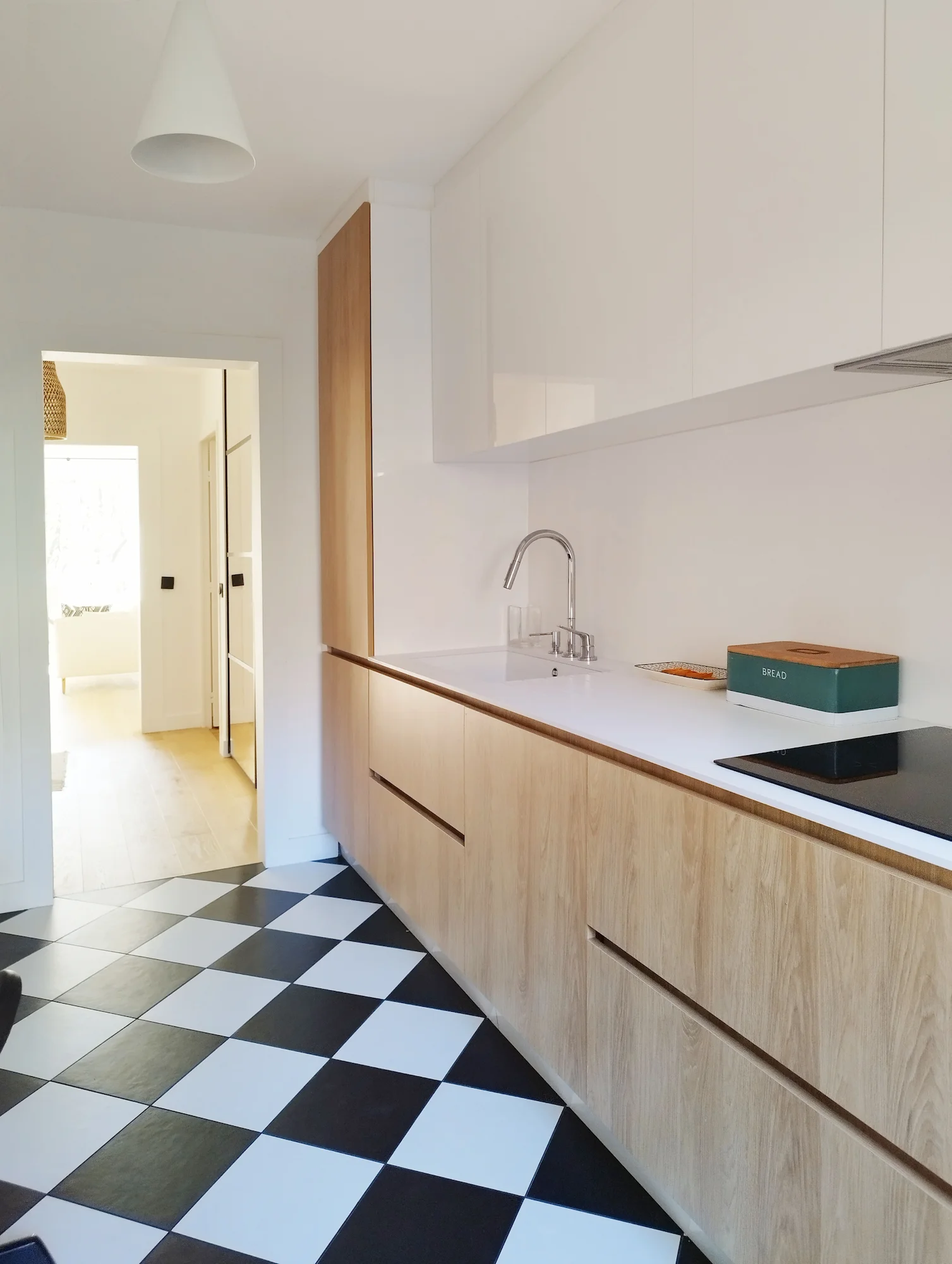 Cuisine linéaire avec rangements en bois et meubles hauts blancs, sol en carrelage damier et circulation ouverte vers le reste de l’appartement.