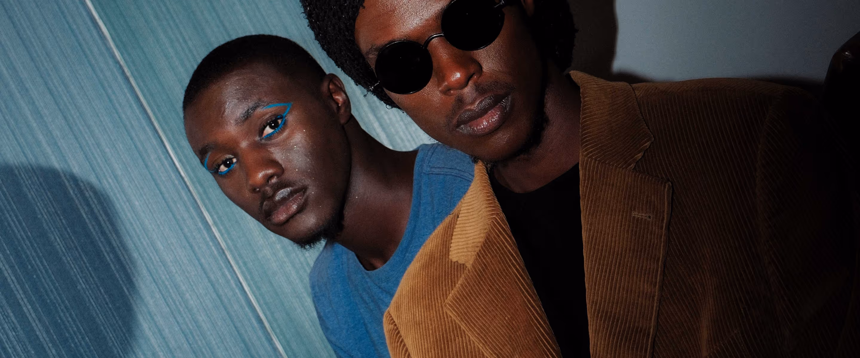 Two young men posing closely together, one wearing blue eyeliner and a blue shirt, the other wearing round black sunglasses and a brown corduroy jacket.