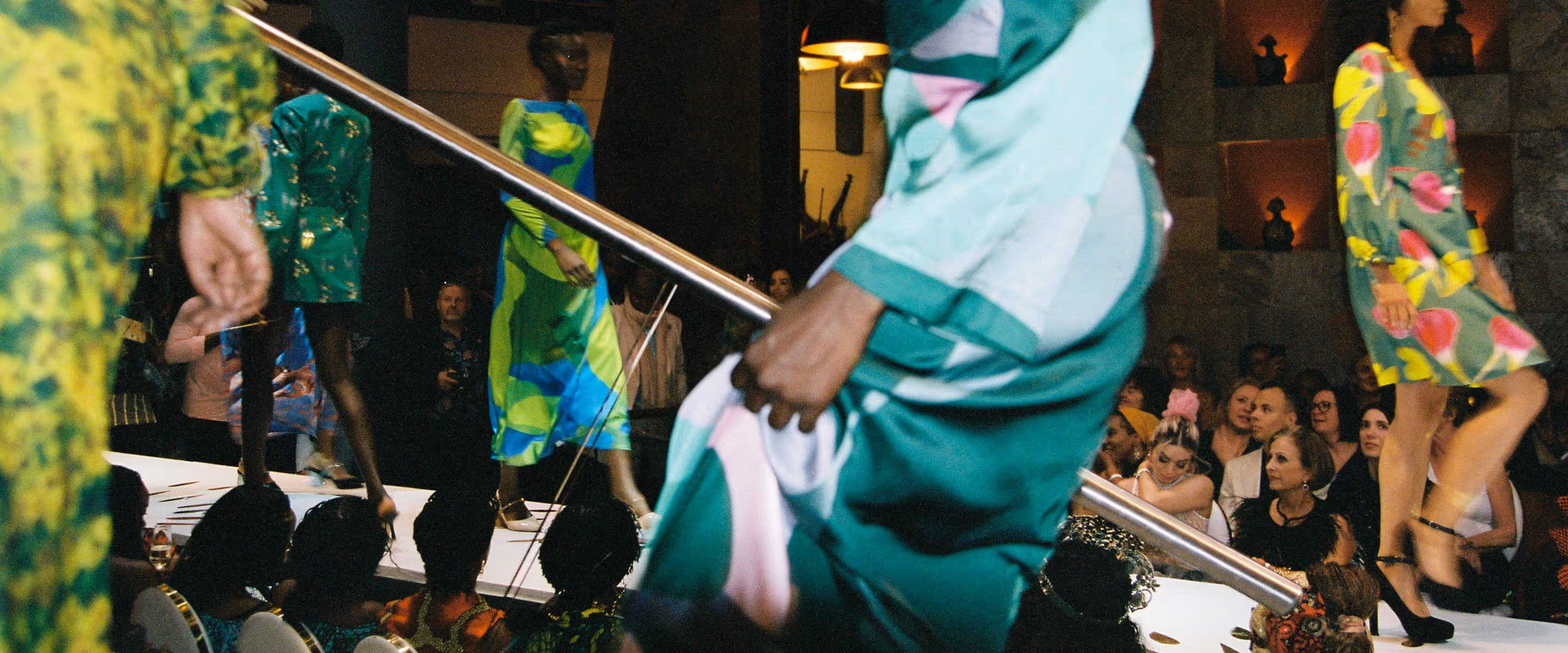 Models walking the runway in colorful dresses during a vibrant fashion show with an attentive audience.