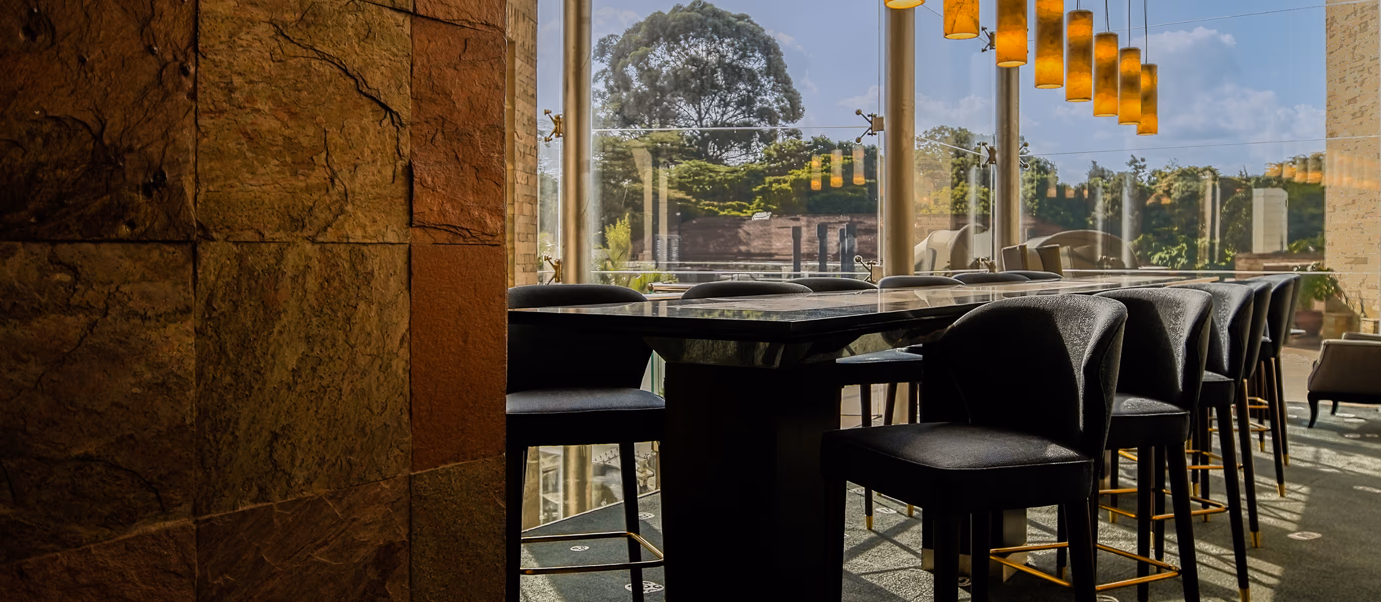 Modern dining area with black high chairs around a dark stone table, large windows, and yellow pendant lights above at Tribe Hotel, Nairobi, Kenya.
