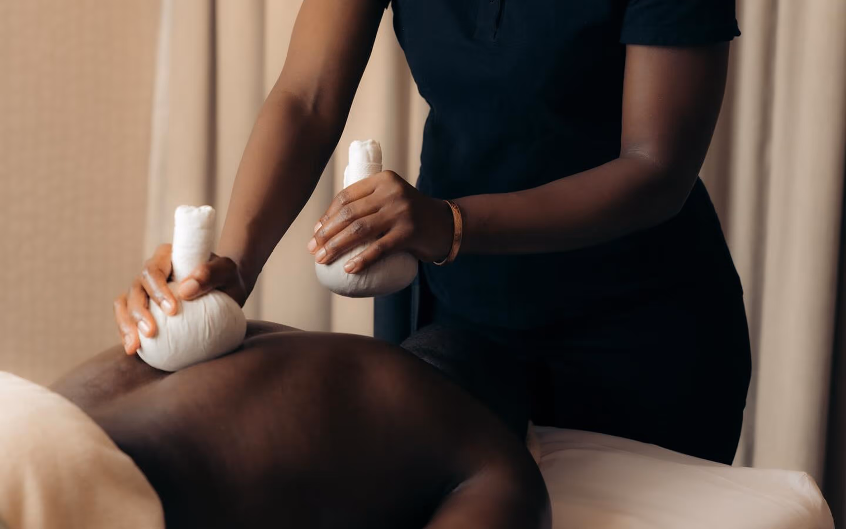 Person receiving a herbal compress massage on their bare back from a therapist wearing black.