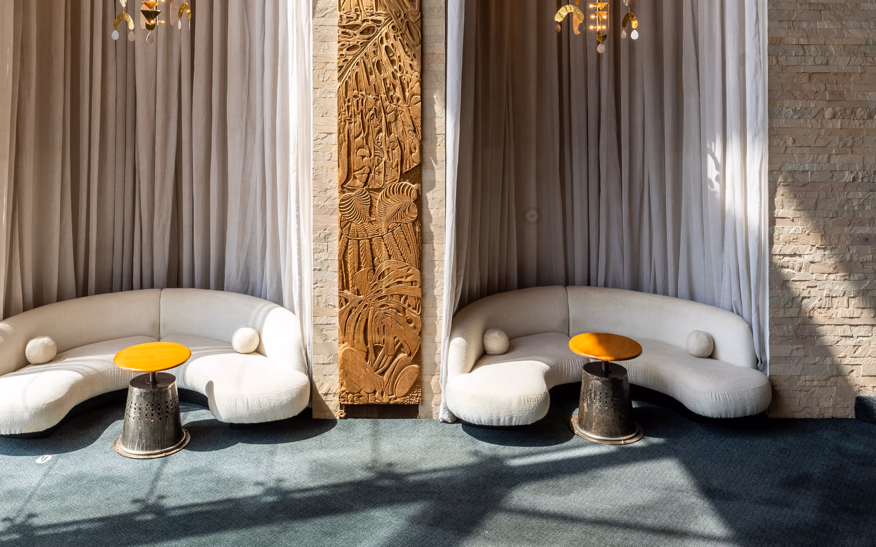 Two white curved sofas with round cushions separated by an ornate vertical wooden carving on a stone wall, each sofa paired with a small round orange table on a blue carpet.