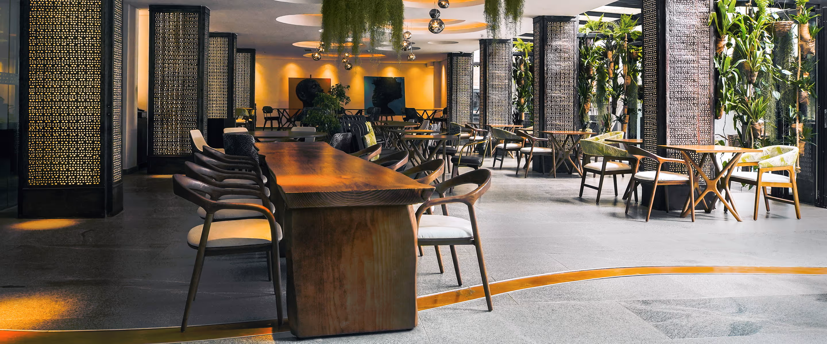 Modern dining area with wooden tables, chairs, hanging plants, and decorative lit panels.