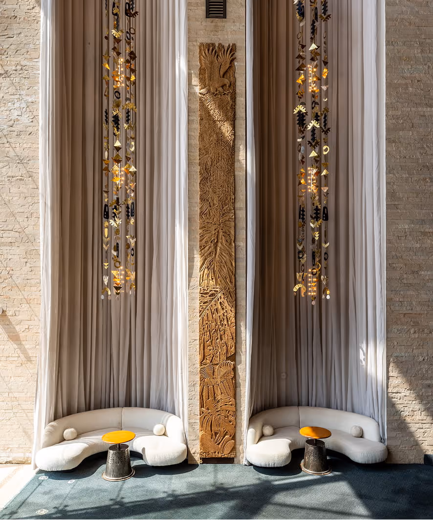 Tribe Hotel in Nairobi, Kenya seating area with two curved white sofas, small orange and grey side tables, long beige curtains, and a tall carved wooden panel between them.