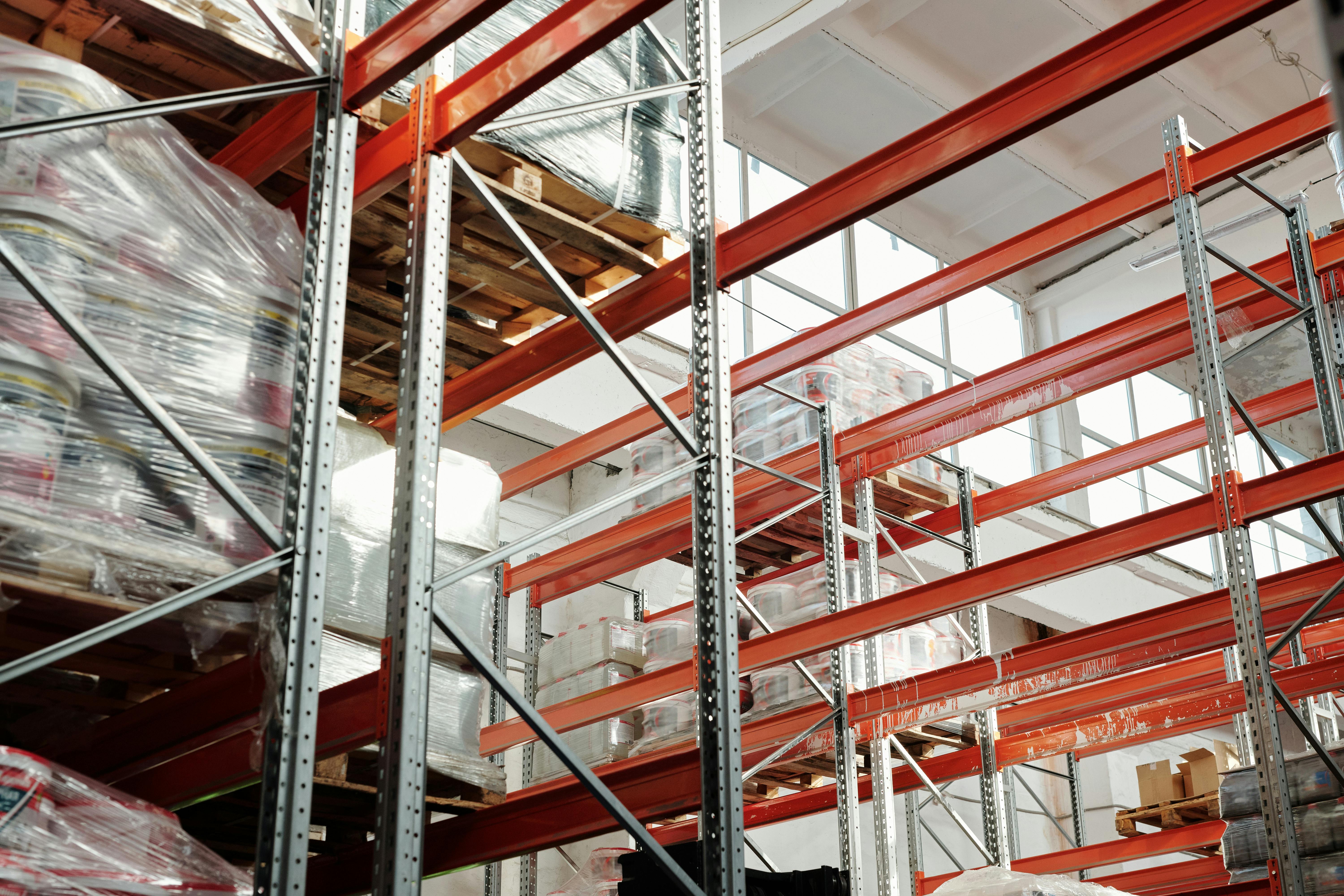 A large commercial warehouse with rows of tall metal shelving units stacked with shrink-wrapped pallets of boxed goods.