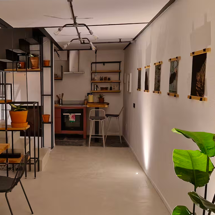 Modern kitchen area with a pink oven, open shelves with kitchen items, and two bar stools, next to a wall with five framed pictures and green plants.