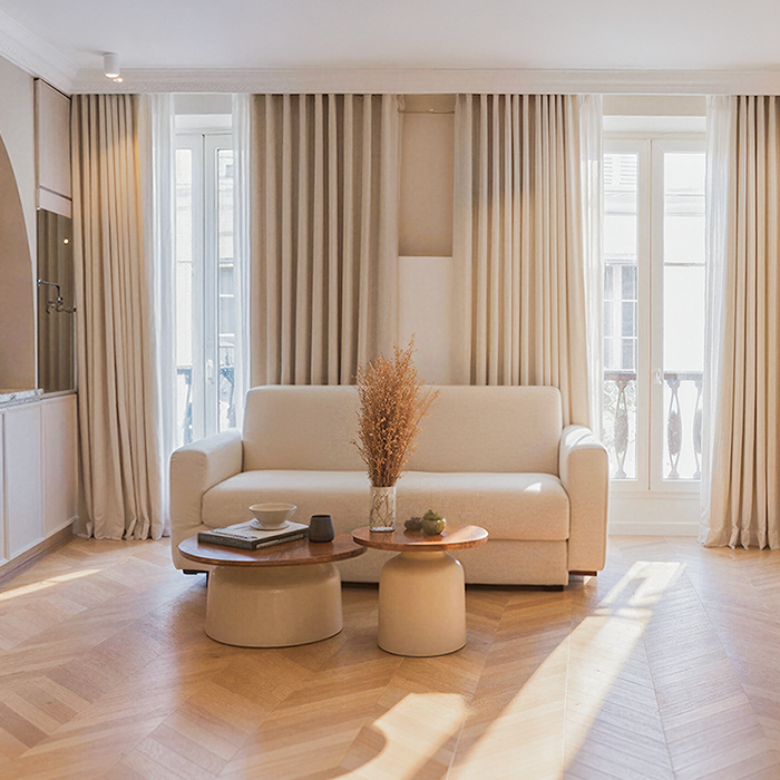 Minimalist living room with beige sofa, two round coffee tables, dried floral centerpiece, and large windows with beige curtains.