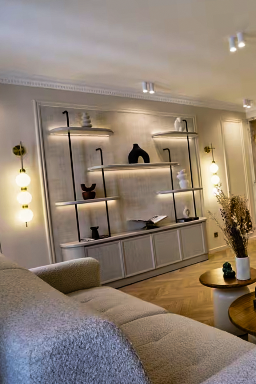 Modern living room with a beige textured sofa, wall-mounted shelves with decorative vases, warm lighting fixtures, and wooden round coffee tables with a white vase of dried flowers.