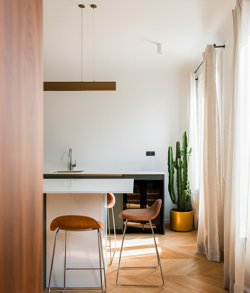 Modern kitchen with two brown bar stools, a white marble island, a tall green cactus in a yellow pot, and sunlight streaming through beige curtains.