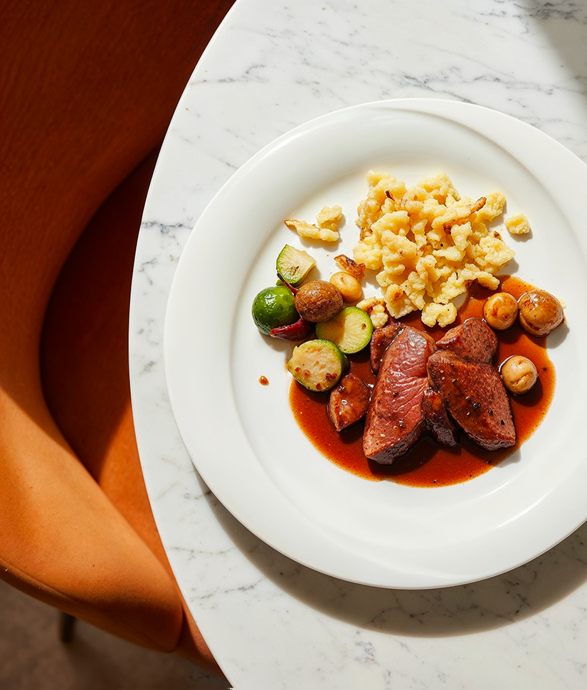 Plate with sliced cooked meat in brown sauce, fried spaetzle, Brussels sprouts, zucchini, and mushrooms on a marble table next to an orange chair.