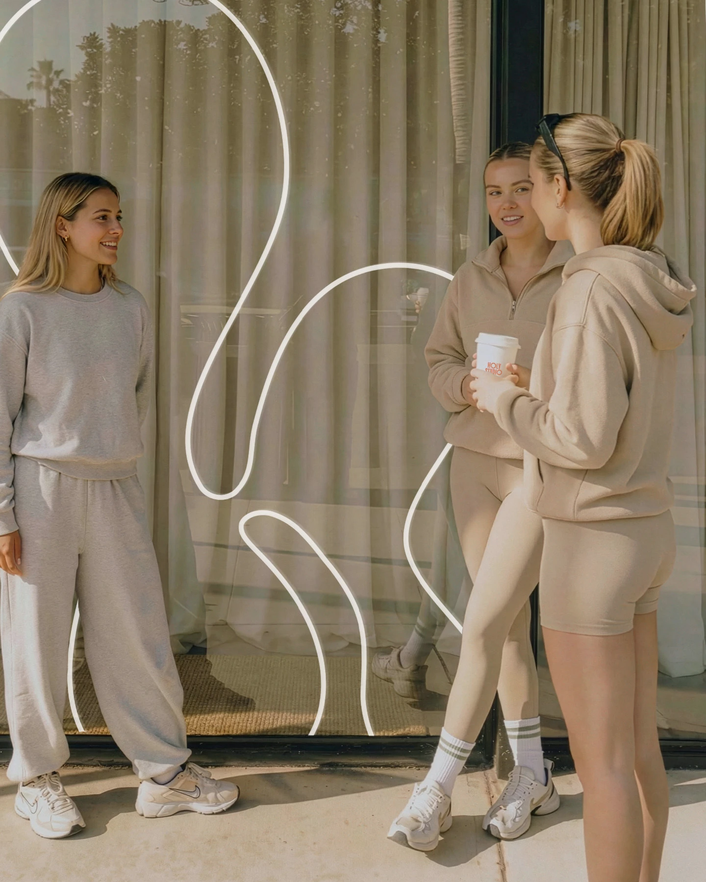 Trzy młode kobiety w sportowych ubraniach rozmawiające na zewnątrz przed oknem z abstrakcyjnym wzorem.