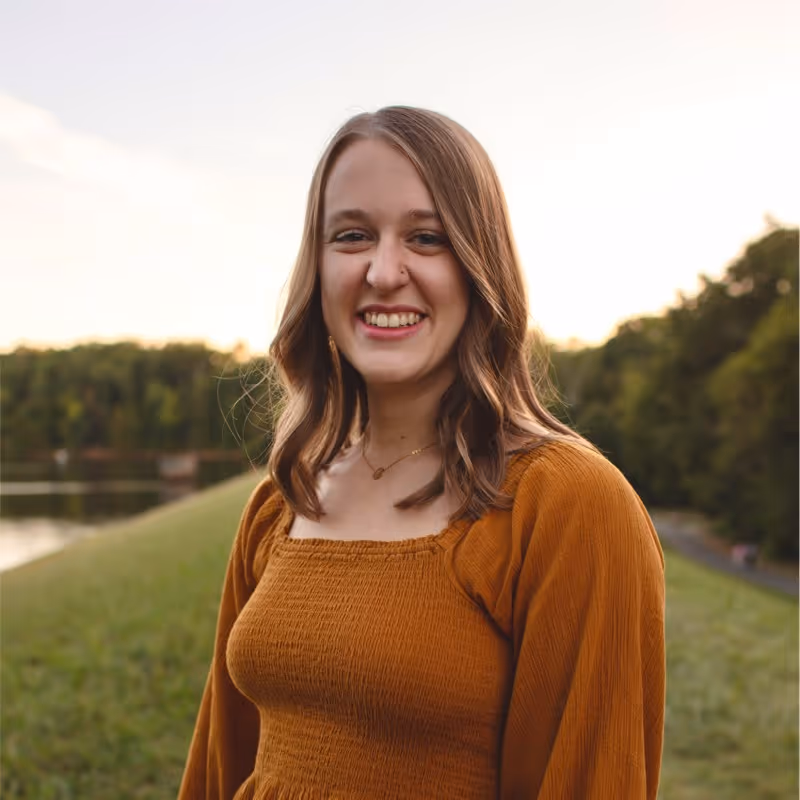 Young woman with shoulder-length wavy hair wearing a rust-colored square-neck top standing outdoors with a blurred background of grass, trees, and water.