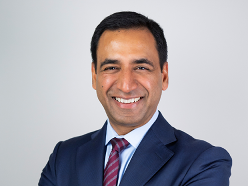 Smiling man in dark blue suit and striped tie with folded arms against a light background.