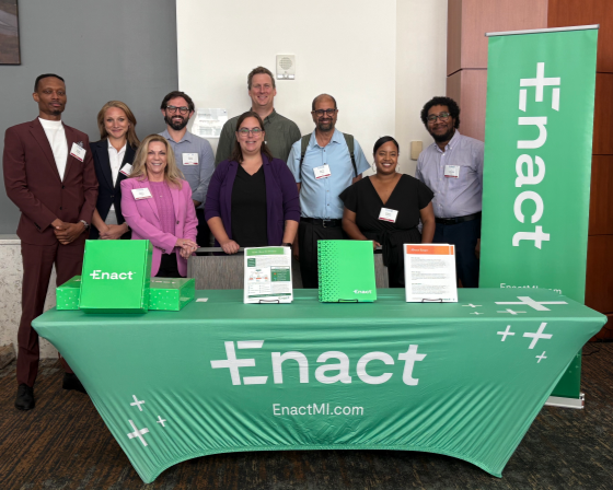 Group of nine diverse professionals standing behind a green table with 'Enact' branding and promotional materials at an indoor event.