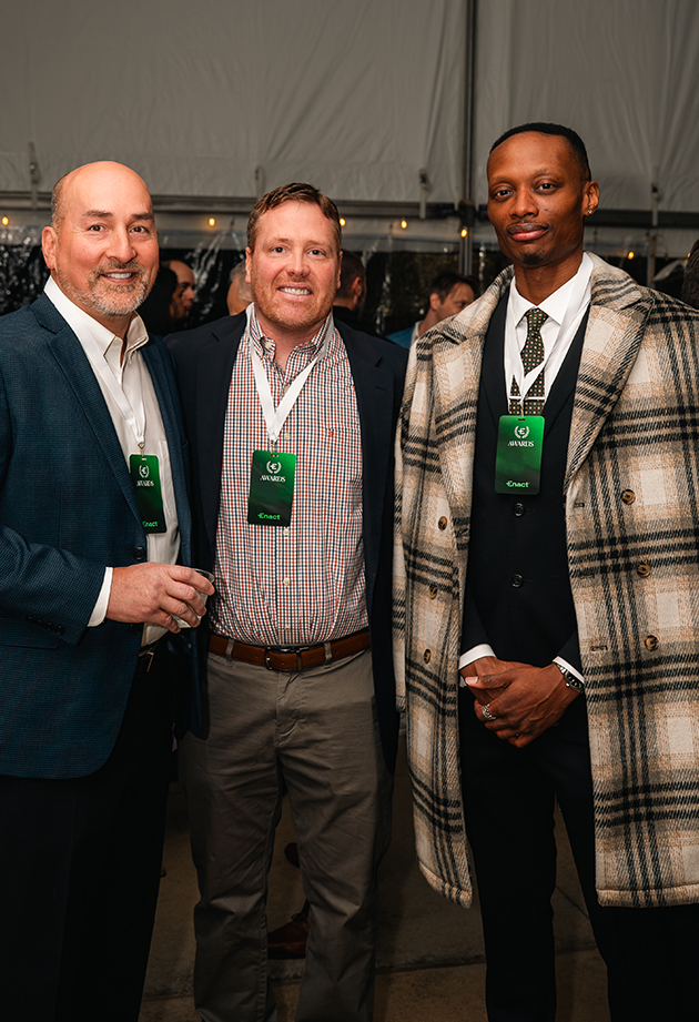 Three men wearing event badges posing together at an indoor gathering under a tent.