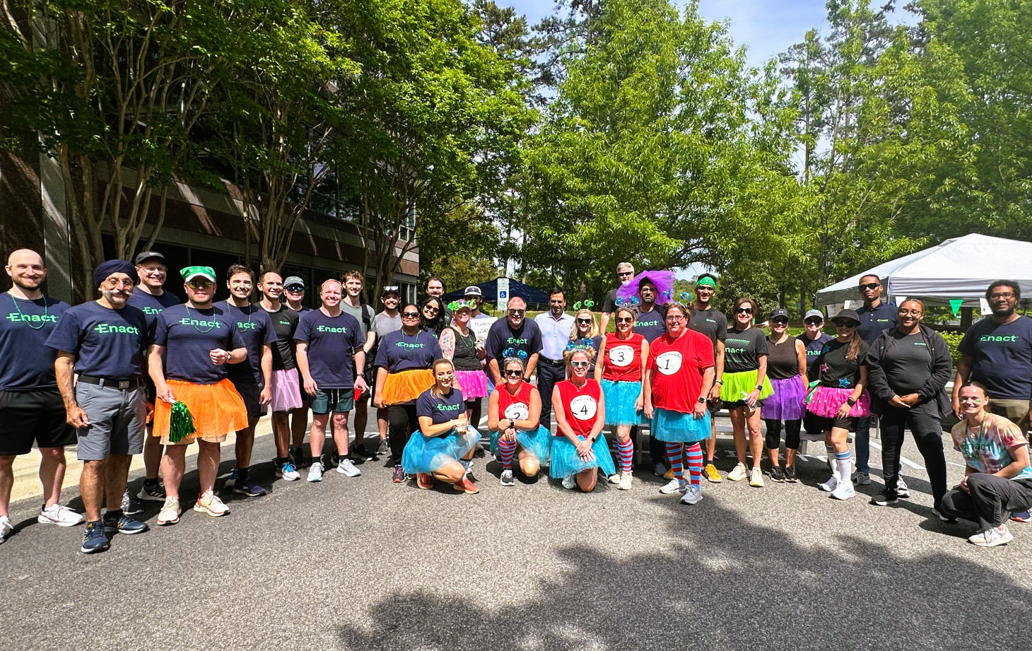 Large group of people outdoors wearing colorful tutus and casual athletic clothes, posing for a group photo under trees after charity run.