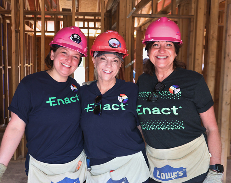 Three smiling women wearing pink construction helmets, Enact t-shirts, and tool belts inside a wooden building frame.