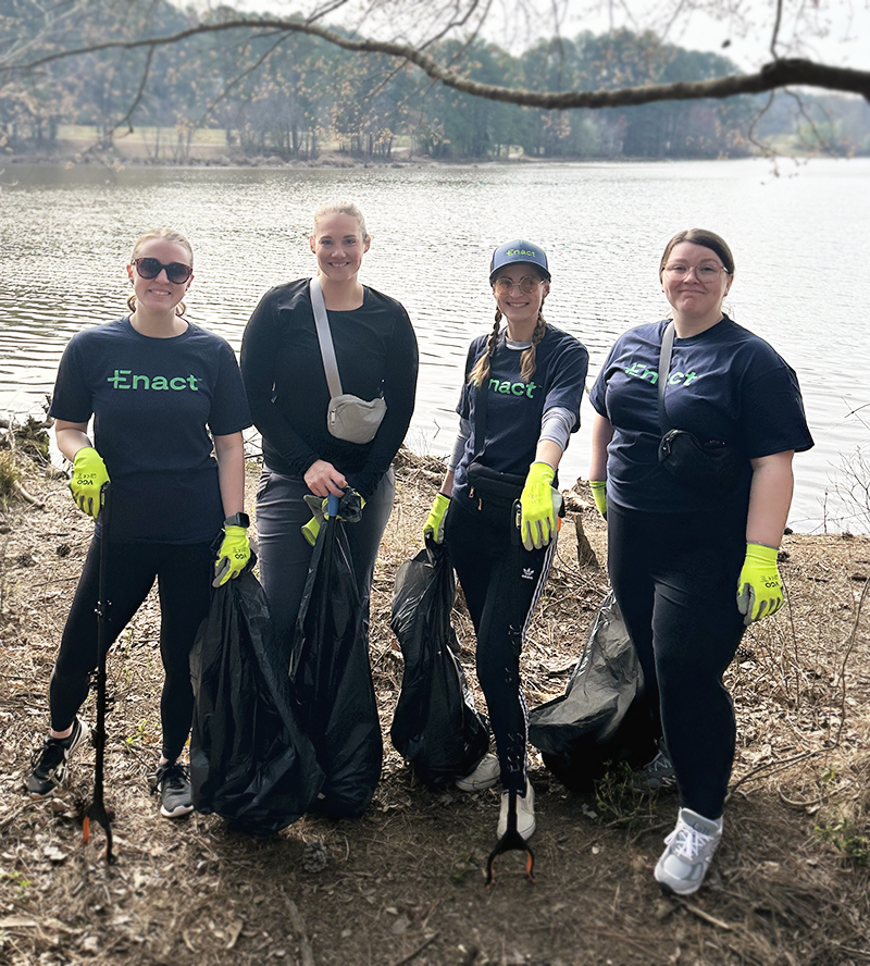 Four people wearing Enact shirts and yellow gloves holding trash bags and litter pickers by a lakeside during a cleanup event.