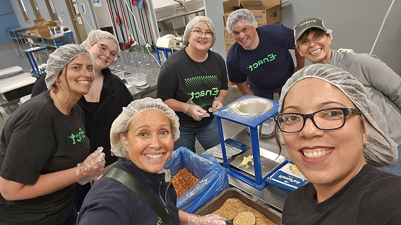 Group of seven smiling volunteers wearing hairnets and gloves, working together in a food packing facility.