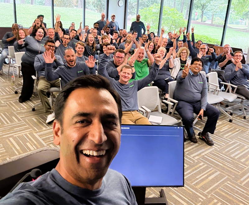 A man takes a smiling selfie with a large group of enthusiastic coworkers wearing the same shirt and seated in a conference room after winning Best Places to Work award. Windows show greenery outside.