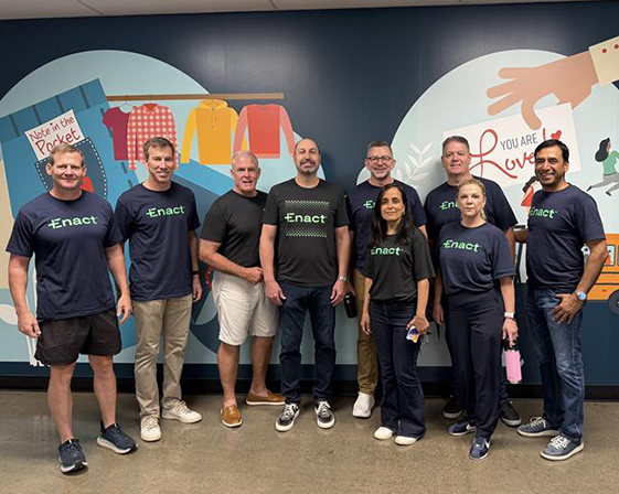 Group of eight people standing indoors in front of a colorful mural, most wearing matching Enact t-shirts.