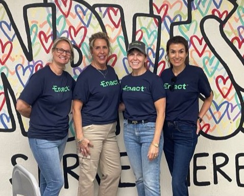 Four women wearing navy Enact t-shirts standing in front of a colorful wall mural with hearts and large letters.