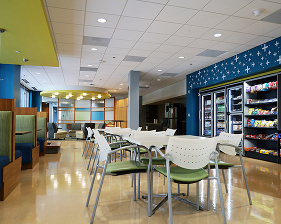 Modern office break room with white tables, green and white chairs, snack shelves, and refrigerated beverages against a blue wall.