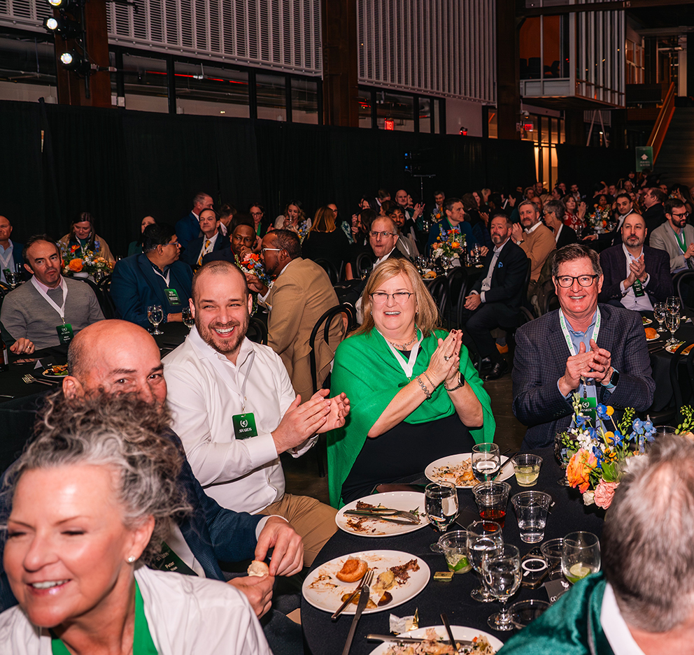 Group of diverse adults seated at round banquet tables, clapping and smiling during an indoor event.