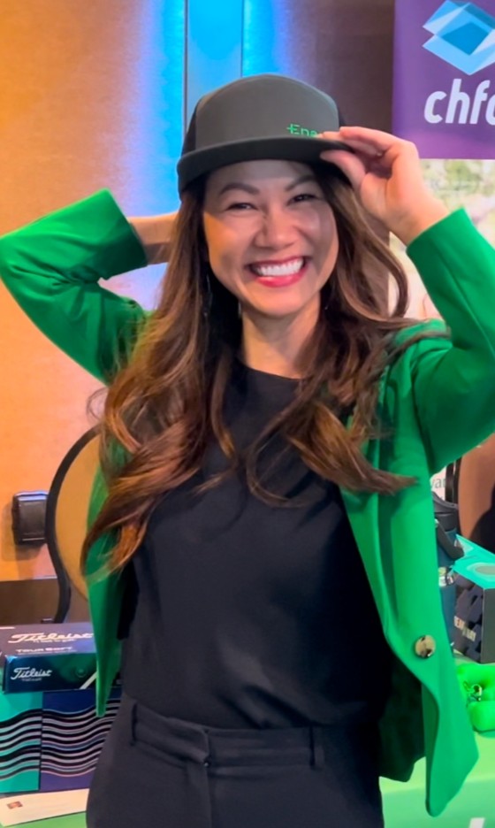 Smiling woman with long brown hair wearing a green blazer and black cap indoors.