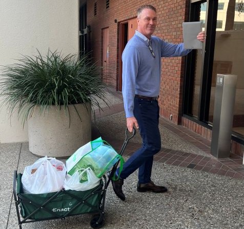 Man in blue sweater and dark pants pulling a green Enact foldable cart loaded with shopping bags near a building entrance.