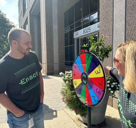 Two people wearing Enact t-shirts smiling and spinning a colorful prize wheel outside an office building.