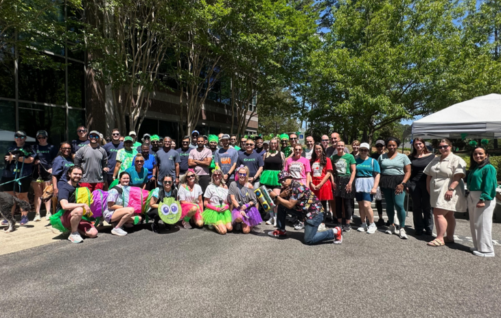 Large group of people outdoors wearing colorful tutus and casual athletic clothes, posing for a group photo under trees after charity run.