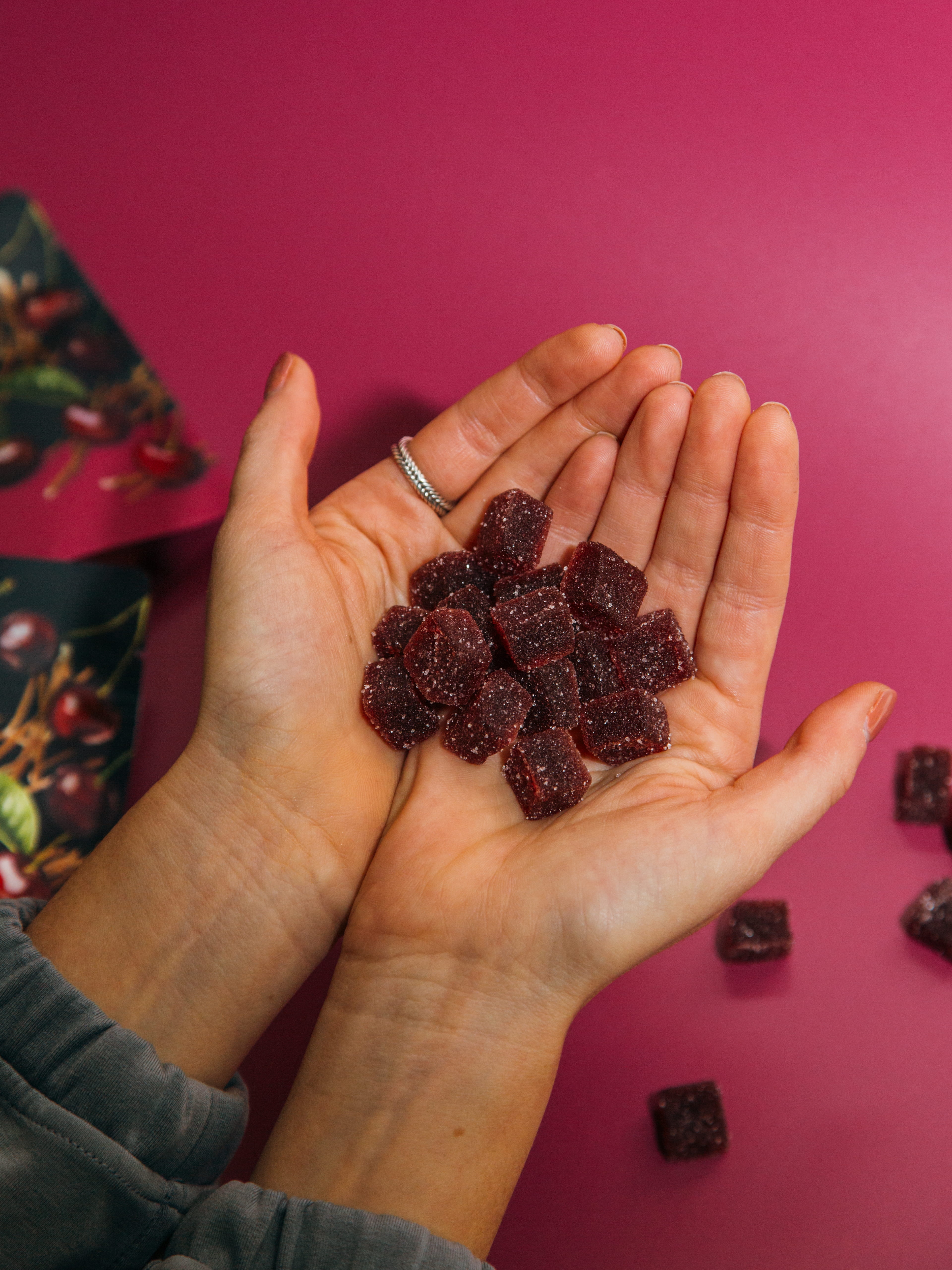 Hands holding several sugar-coated dark red gummy candies against a pink background.