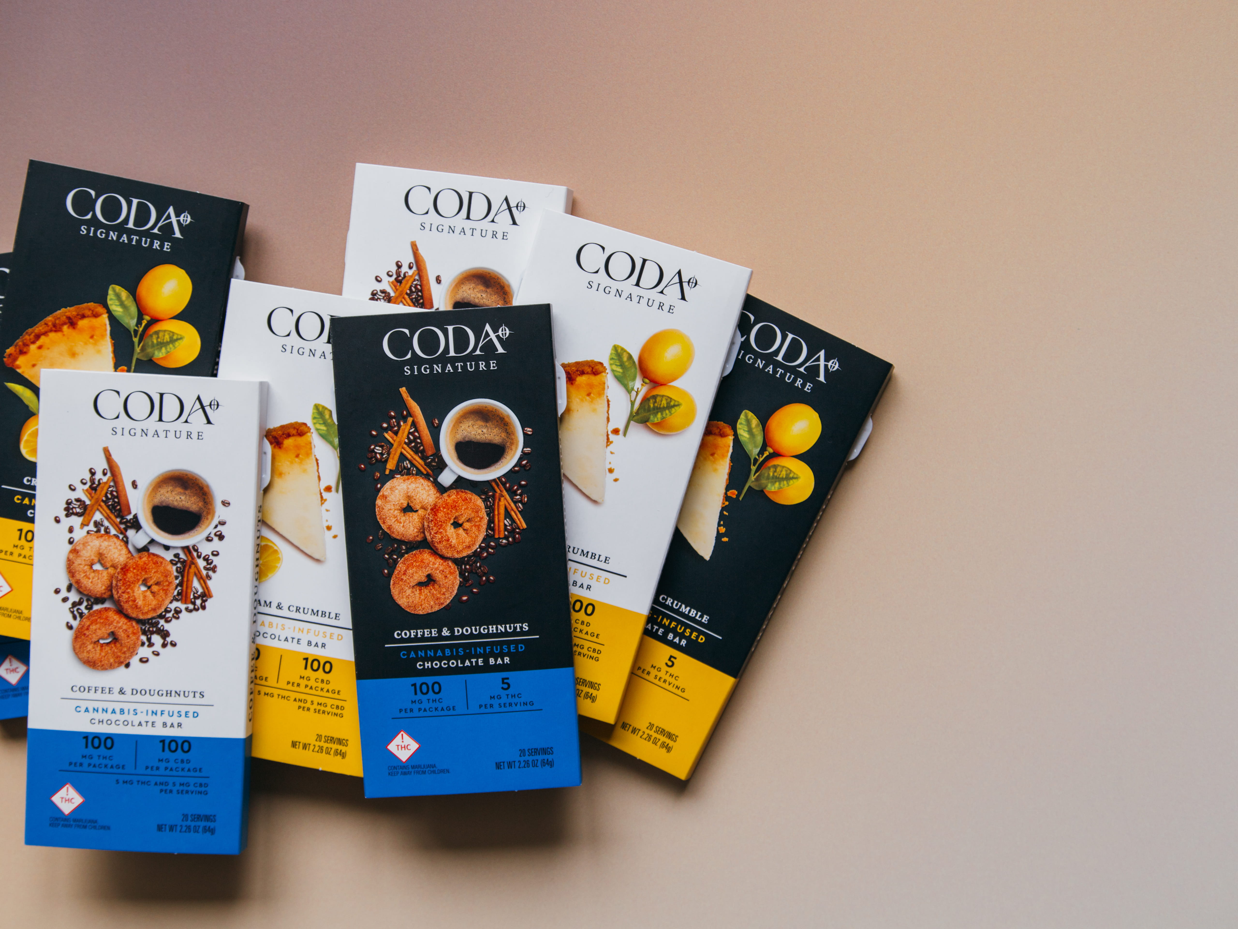 Assorted CODA Signature cannabis-infused chocolate bars in Coffee & Doughnuts and Cream & Crumble flavors on a beige surface.